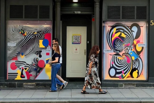 People walk past a storefront with colorful, abstract art displays on both windows.