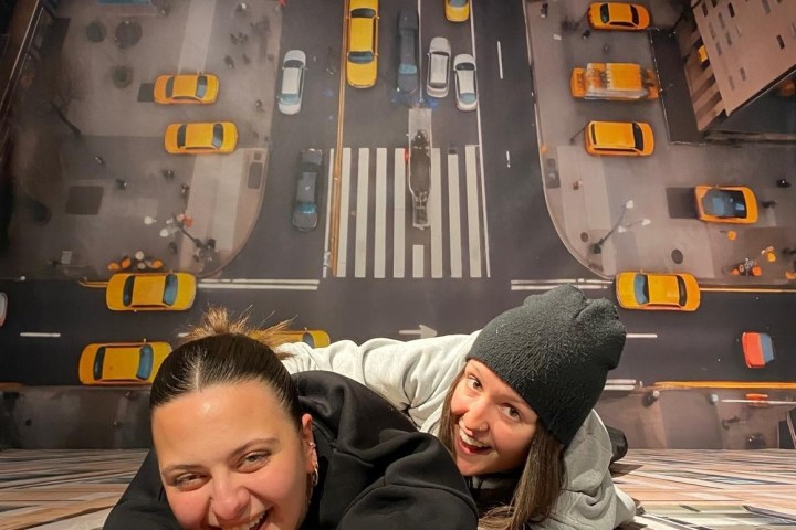 Two people on a floor mural of a city street with taxis, appearing to hang from a building.